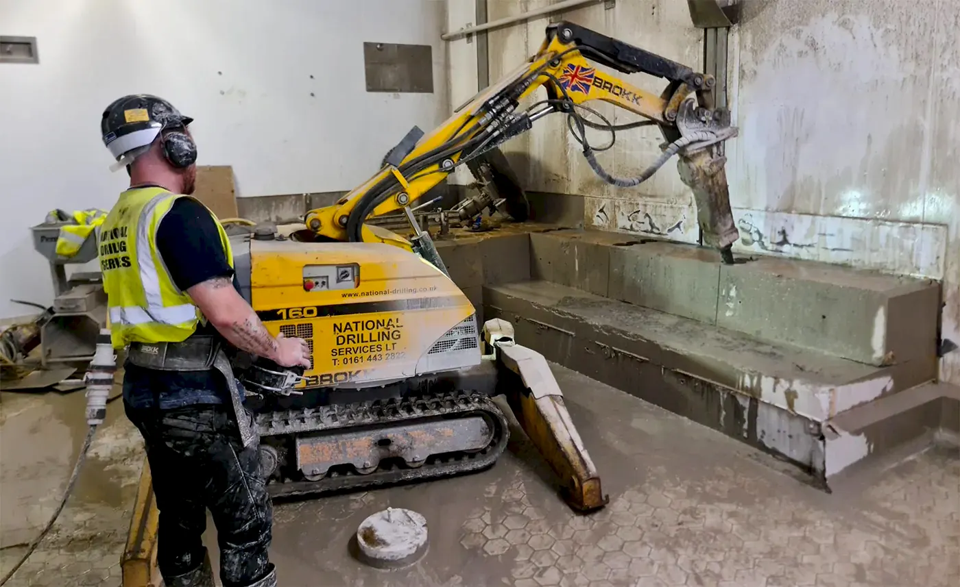 Robotic demolition machine operated by worker, featuring National Drilling Services branding, in a construction site setting with concrete surfaces and tools.