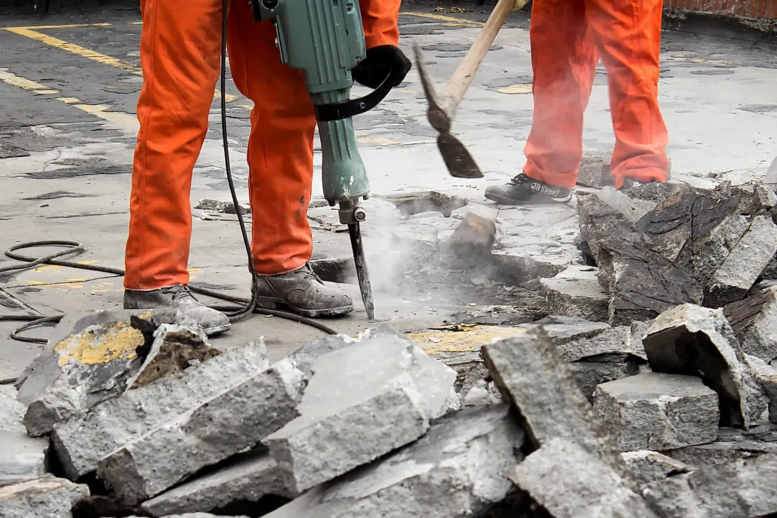Construction workers using a jackhammer on concrete surface with broken concrete debris around, showcasing diamond core drilling services by National Drilling Services.