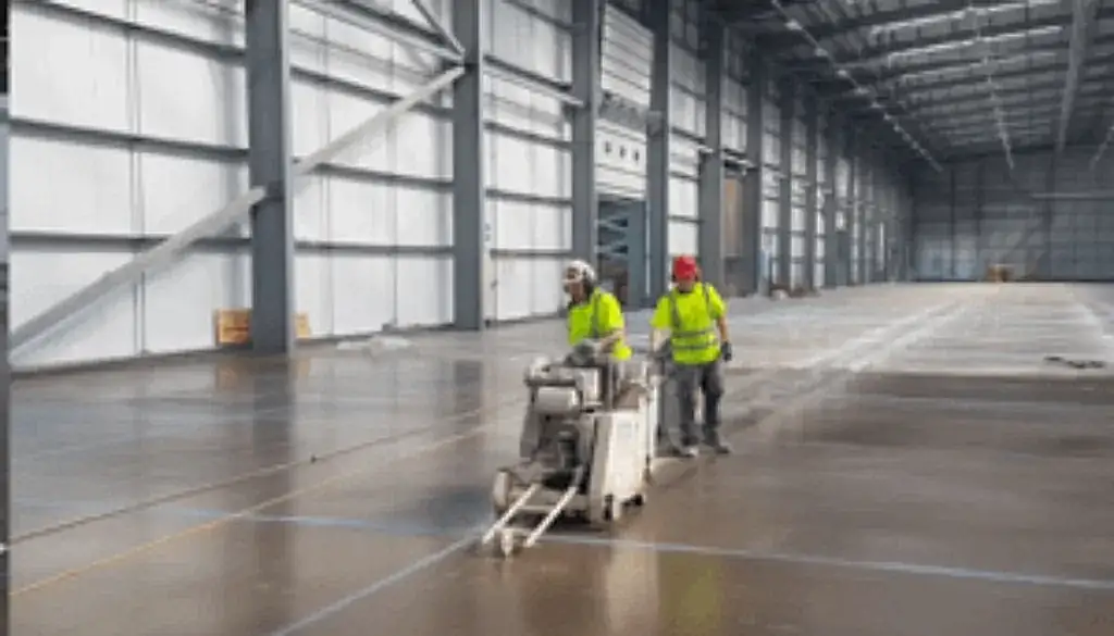 Construction workers operating a concrete floor saw in a spacious industrial setting, emphasizing eco-friendly concrete sawing services by National Drilling Services.