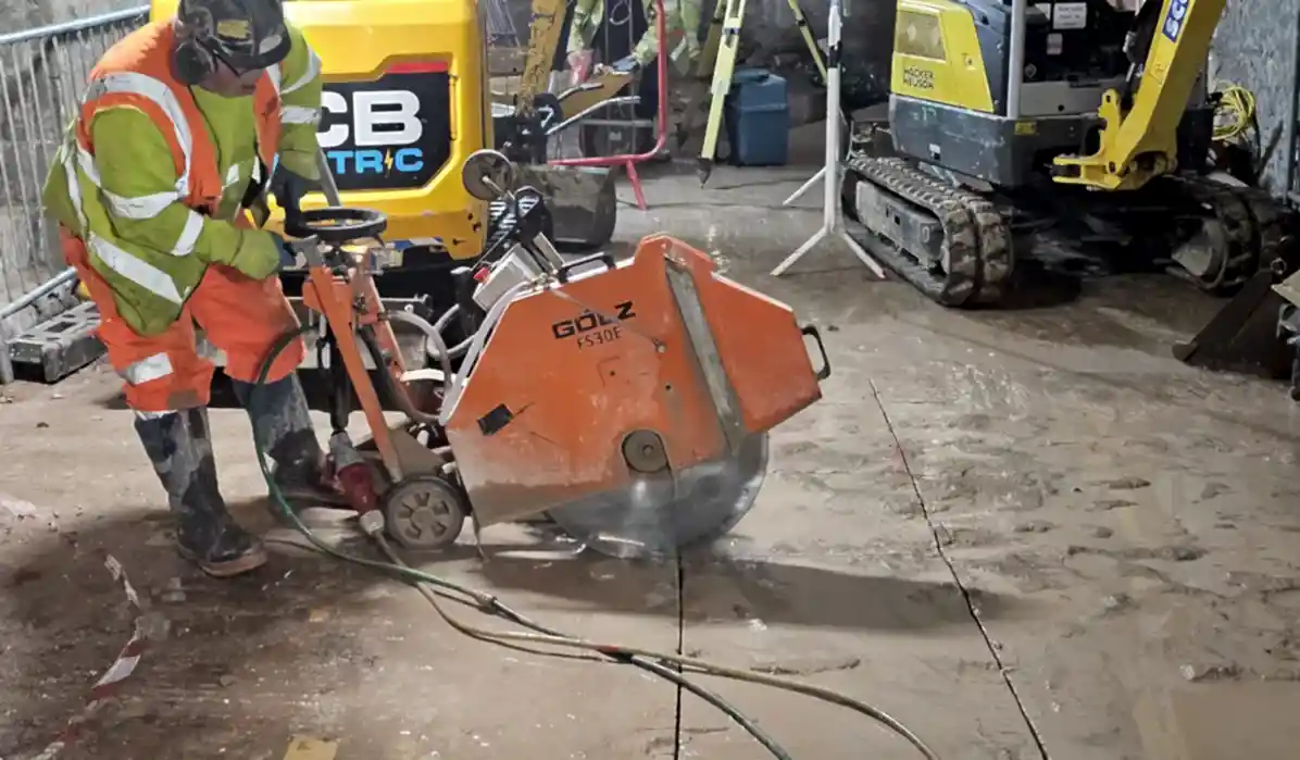 Construction worker operating an electric floor saw on a concrete surface, surrounded by construction machinery and safety barriers, illustrating concrete floor sawing services by National Drilling Services.