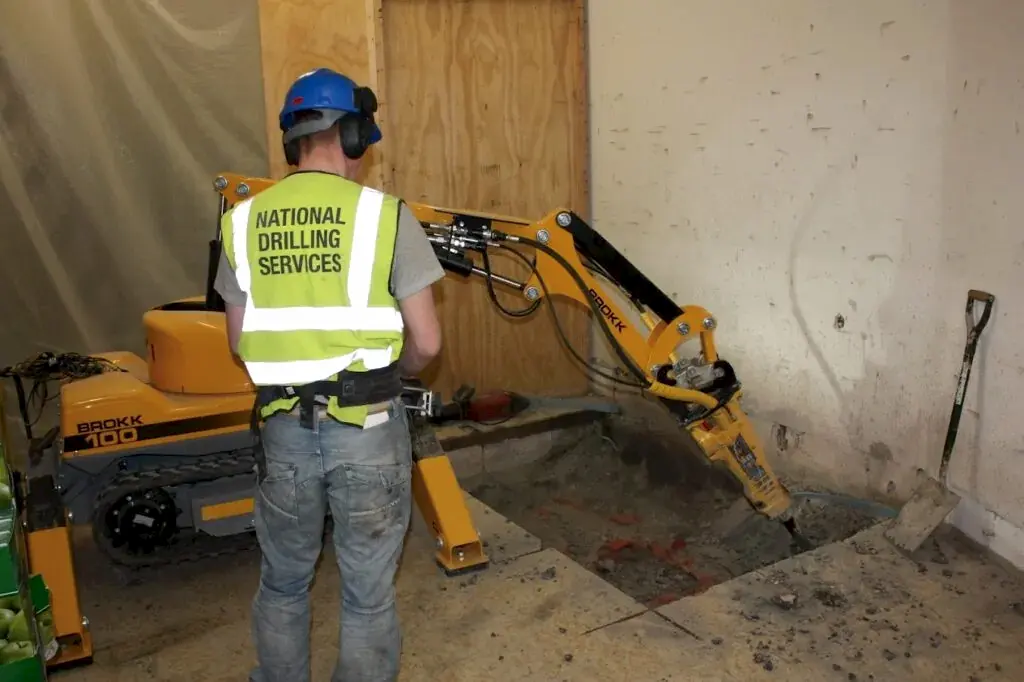 Worker operating a Brokk 100 robotic demolition machine with a hydraulic attachment, wearing a safety vest marked "National Drilling Services," on a construction site in Manchester.