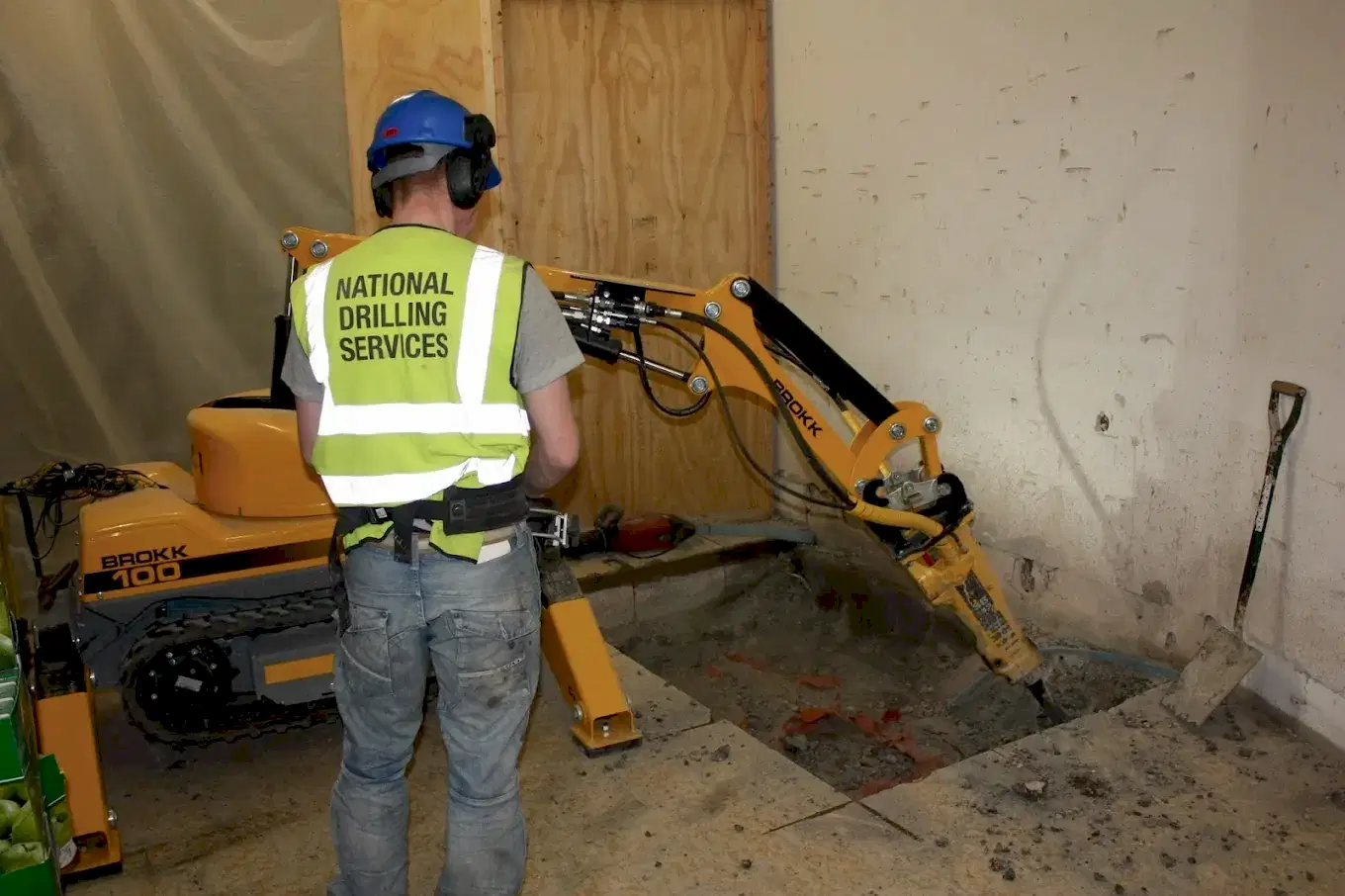 Worker operating a Brokk robotic demolition machine indoors, wearing a safety vest with "National Drilling Services," showcasing advanced concrete contracting technology.