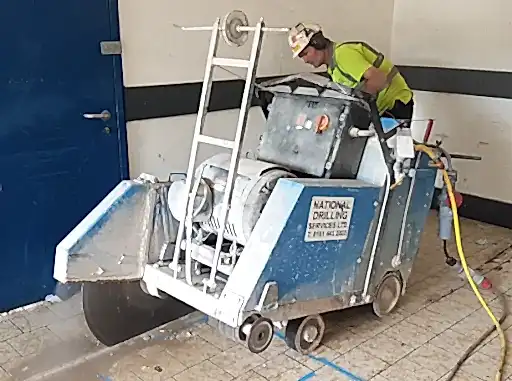 Concrete sawing machine operated by a technician from National Drilling Services, cutting through a concrete floor in an indoor setting.