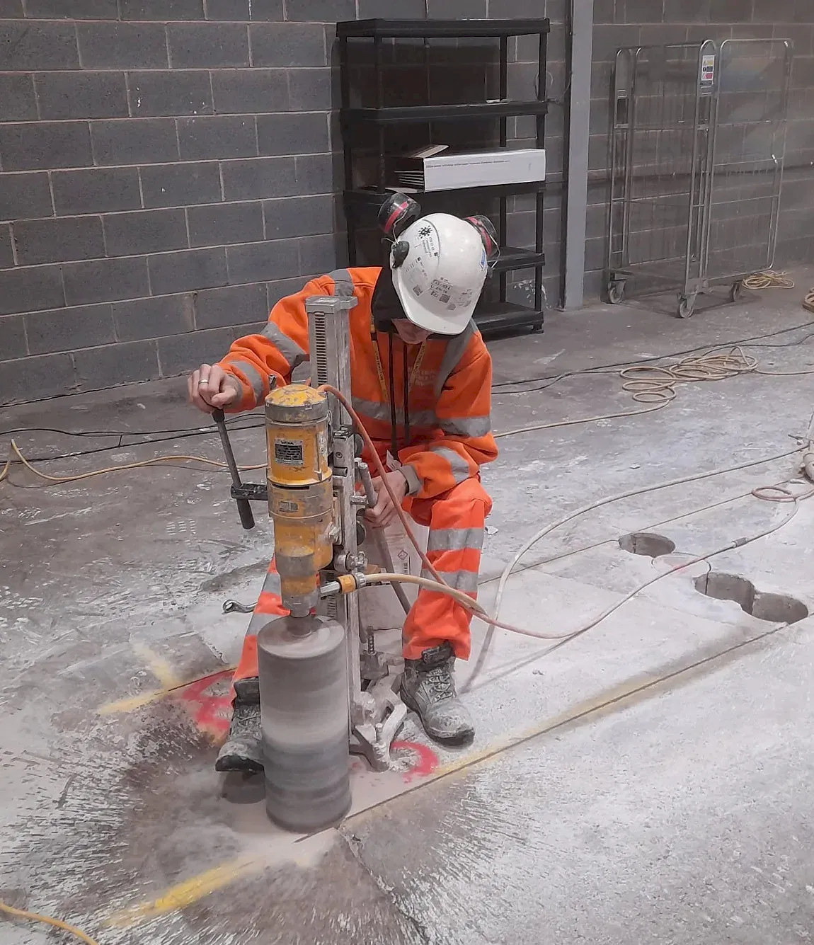 Worker operating diamond core drilling machine on construction site, wearing safety gear, with visible core samples and dust.