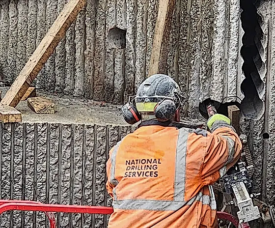 Worker from National Drilling Services operating diamond core drilling equipment on a concrete wall, showcasing precision cutting techniques in construction.