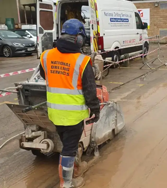 Worker in high-visibility vest with "National Drilling Services" logo operating a concrete floor saw on a construction site, with a company van in the background.